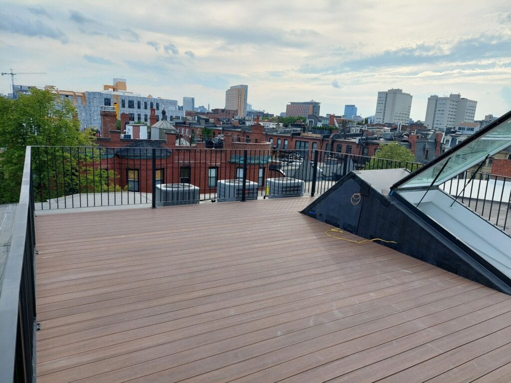 Rooftop terrace with wooden decking, custom iron work detailing around the glass skylight, and city skyline in the background under a cloudy sky.