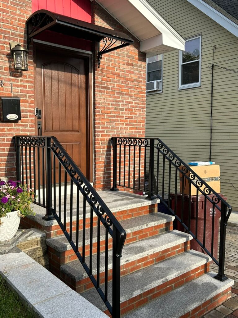 A brick house entrance features a brown door and black metal railing, likely crafted by a skilled contractor. A small overhang shelters the steps, which are adorned with potted flowers and a package on the right side.