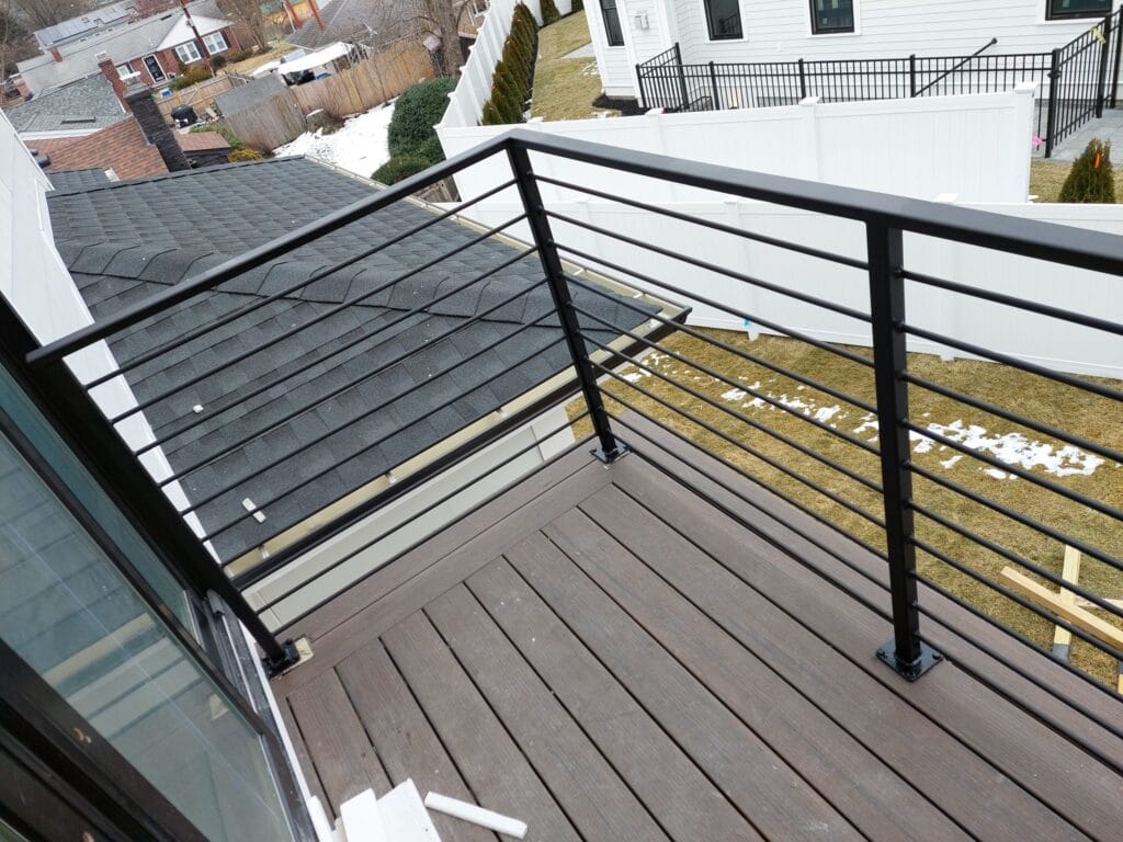 A small balcony with custom iron work railings overlooks a backyard. The view includes neighboring houses, a roof, and patches of snow on the grass.