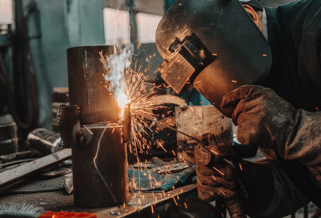 A welder in protective gear expertly works on a metal piece, with bright sparks flying throughout the workshop, showcasing skilled wrought iron work.