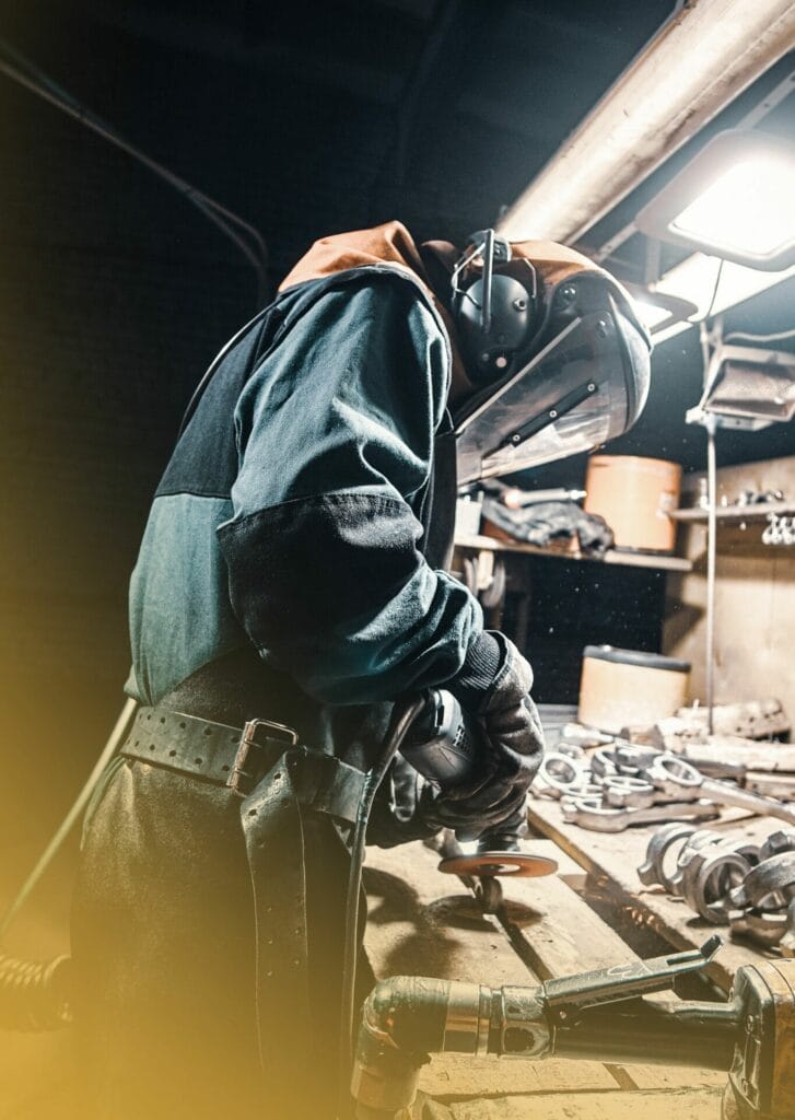 Person in safety gear using a power tool while working on iron work repair at a cluttered workbench in a workshop.