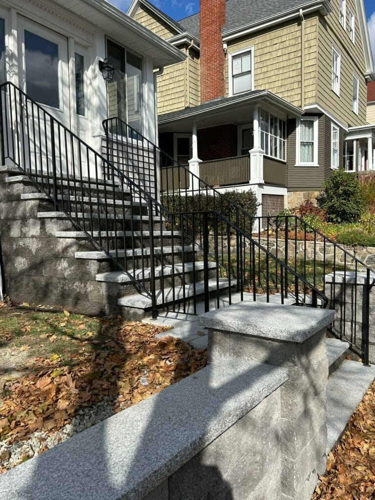 Stone steps with black railings leading to a house with a porch. Steps are bordered by a stone wall. Fallen leaves on the ground.