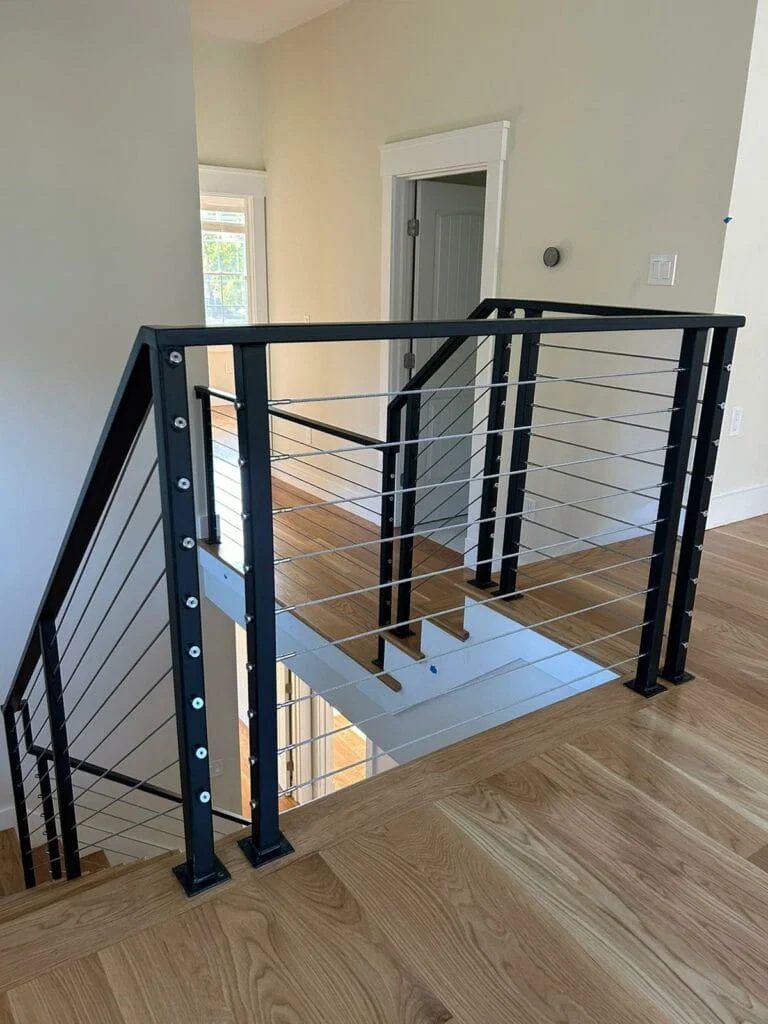 Black metal railing with cable wires on a wooden staircase landing in a modern interior space.