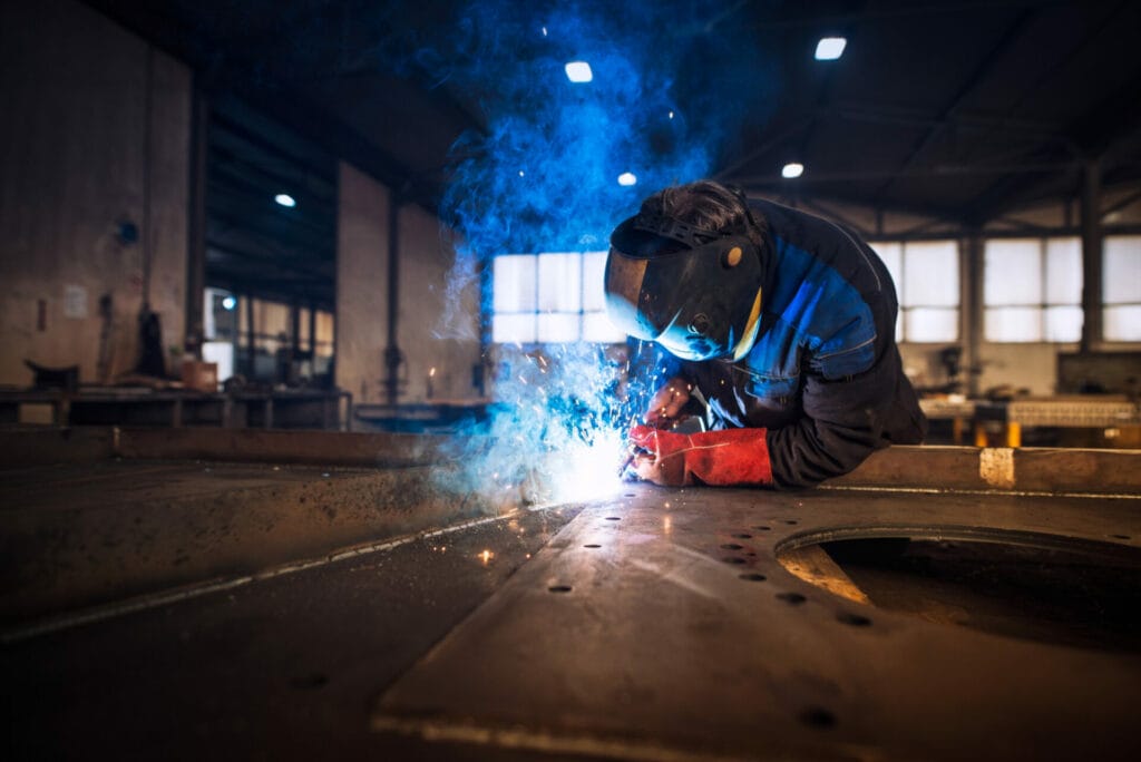 A person in safety gear welding metal in a factory, with sparks and blue smoke rising around them, showcases skilled metal work in action.