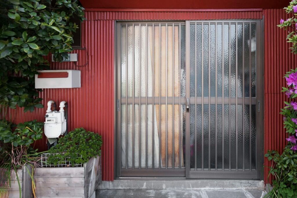 Red house exterior with metal sliding door, security grilles, utility meters, plants, mailbox, and purple flowers on the right.